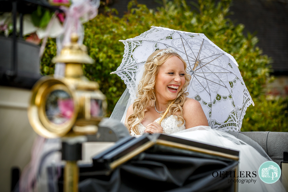 Happy Bride in an open top carriage laughing under an umbrella shielding herself from the sun.