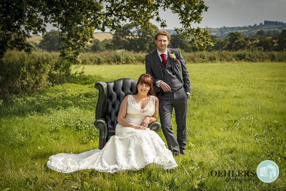 Bride sitting on an armchair whilst grrom leans on it behind in the beautiful countryside.