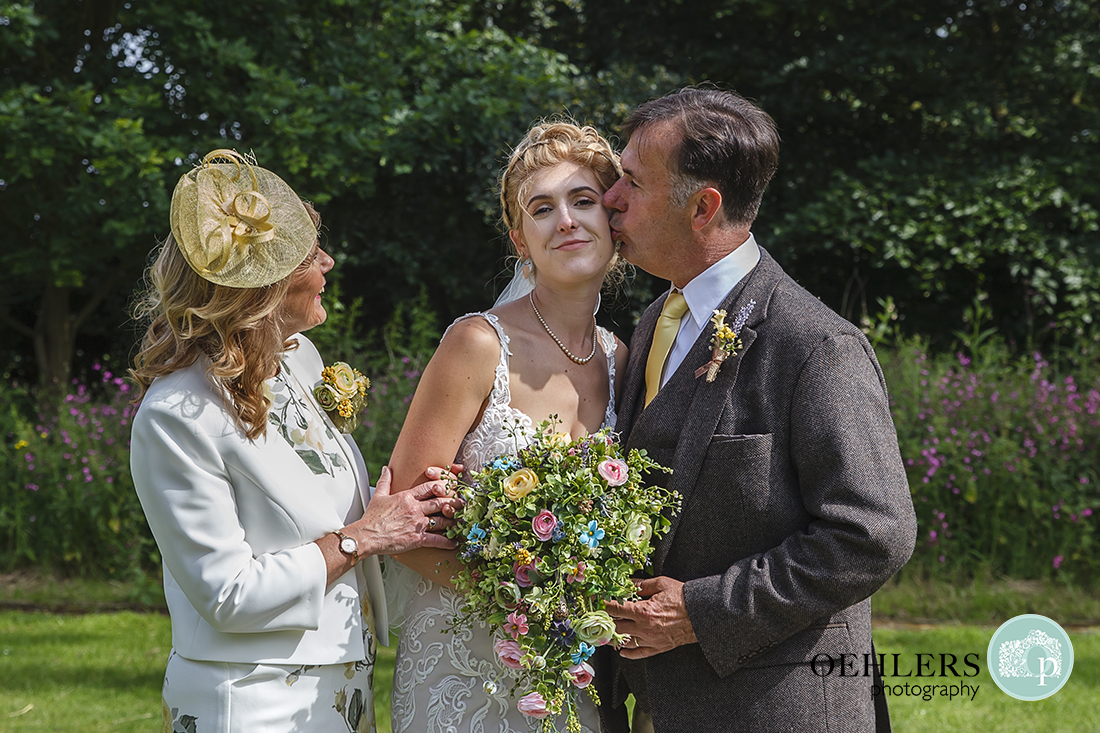 Brides parents with dad kissing the bride on her cheek