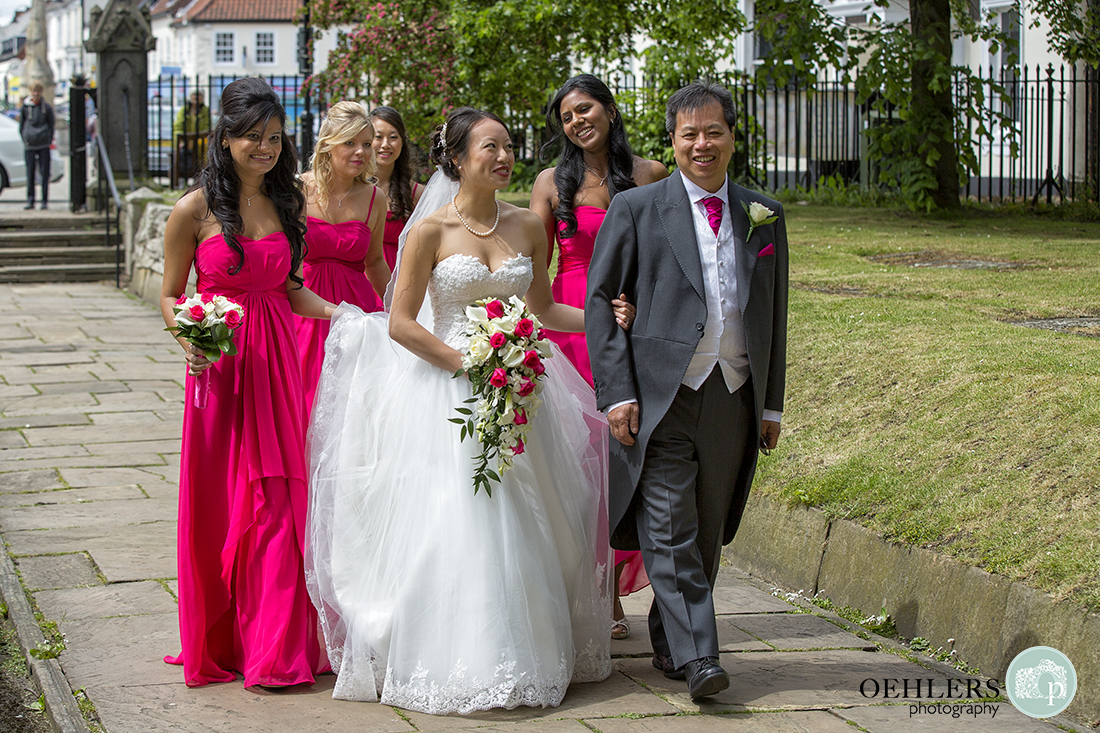 Bride and father walking arm in arm down the path leading to Selby Abbey with Bridesmaids holding the train.