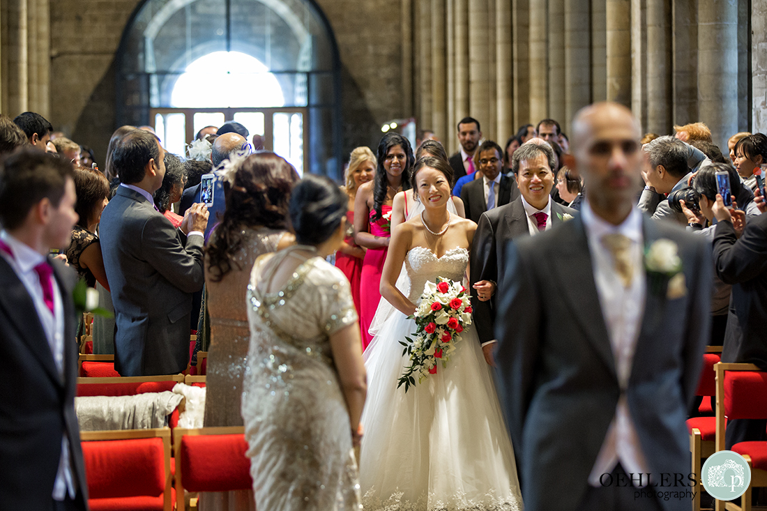 Bride looking at the groom's mother as she walks down the aisle with her father and bridesmaids whilst the groom waits in anticipation.