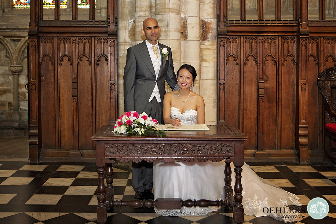 Portrait of the Bride sitting down and the Groom standing whilst the bride pretends to sign the register.