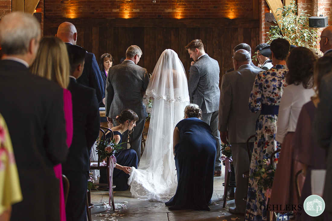 Shustoke Barn Wedding Photographers-Two bridesmaids arranging the back of bride's dress before the start of the ceremony as guests look on.