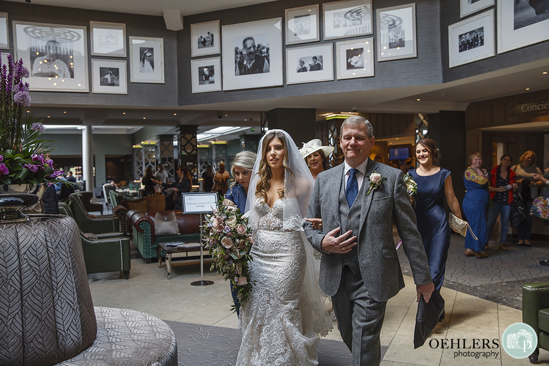 Bride and father arm in arm walking through the rotunda at The Belfry Golf Course with mother and bridesmaids behind.