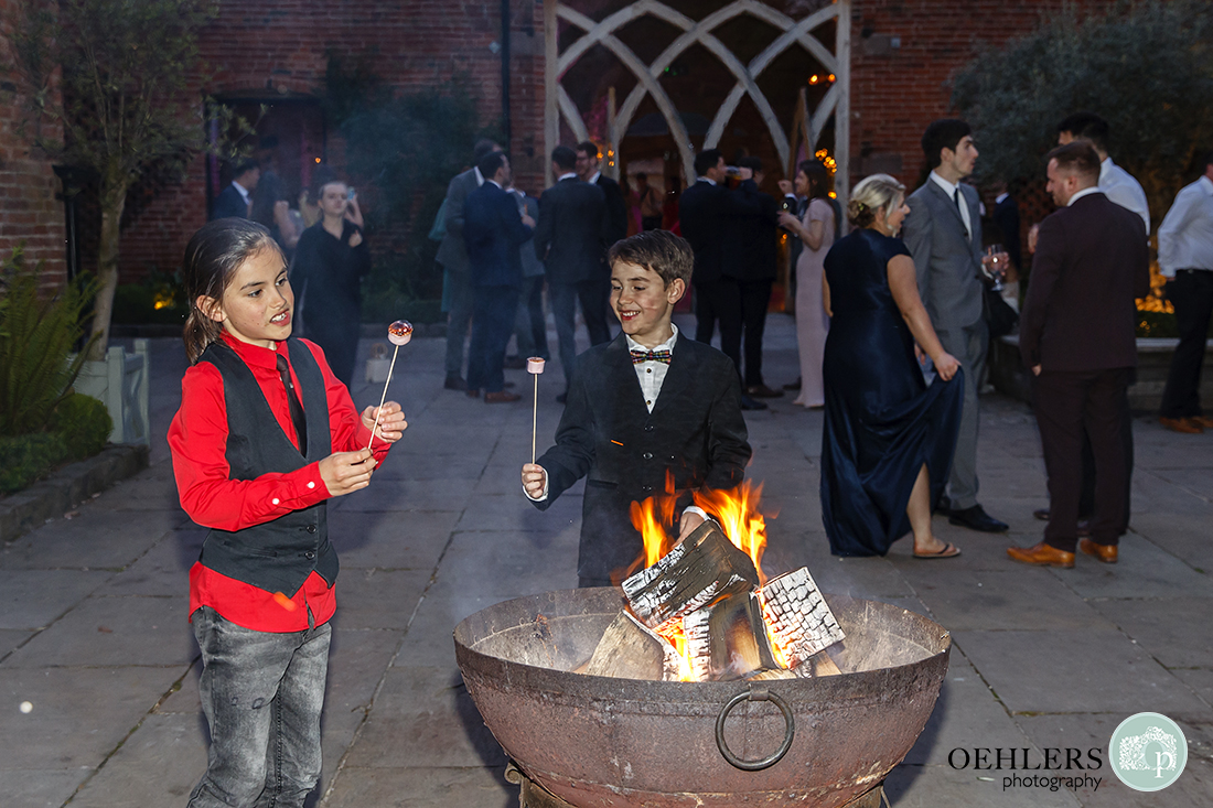 Shustoke Barn Wedding Photographers-Two young guests enjoying toasting marshmallows over a fire pit in the courtyard in the evening.