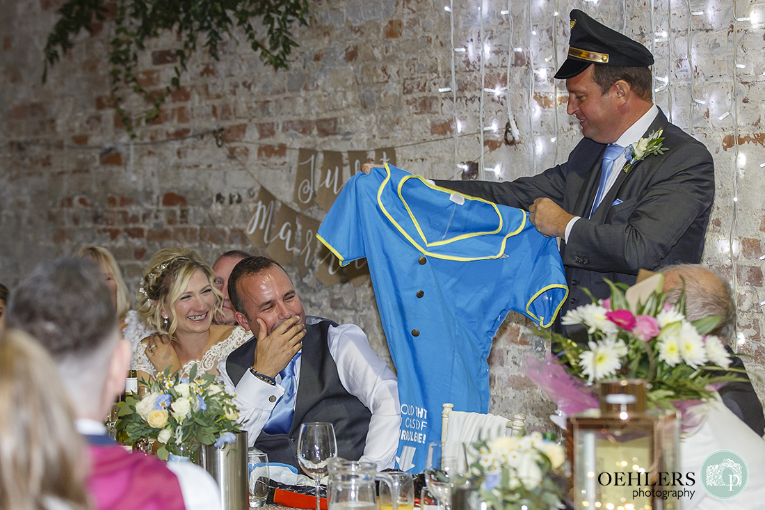 groom and bride laughs at bestman's speech as he holds up outfit the groom wore for his stag do.