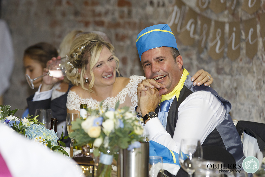 bride puts her arm around embarrassed groom wearing an air hostess hat during the speeches