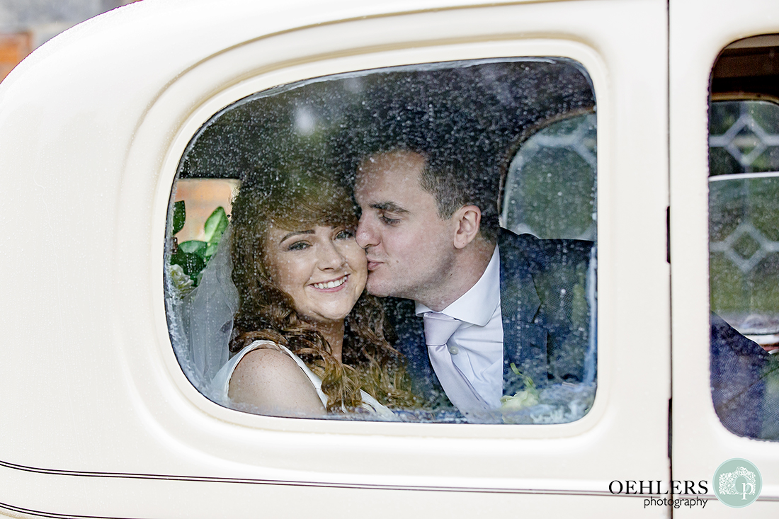 The groom kisses his bride behind a rain droplet window of their wedding car.