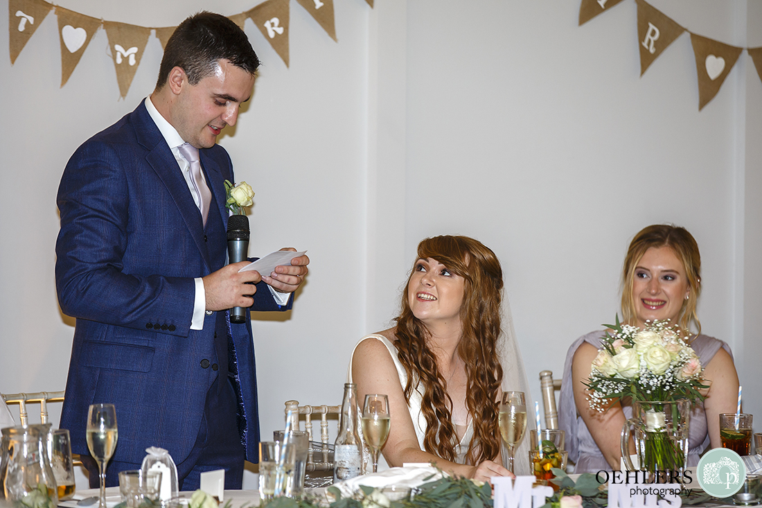 Groom standing up at the top table whilst the bride looks at him.