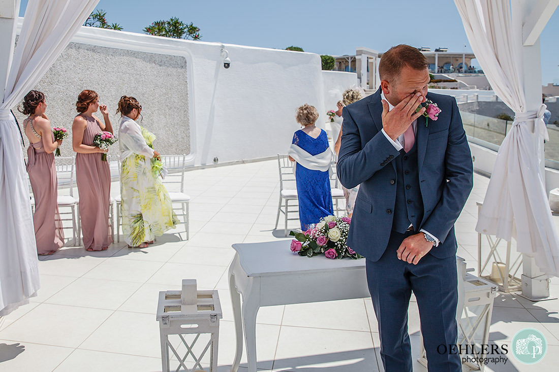 Santorini Destination Wedding Photographers - after spotting his bride, the groom emotionally wipes a tear from his eyes.