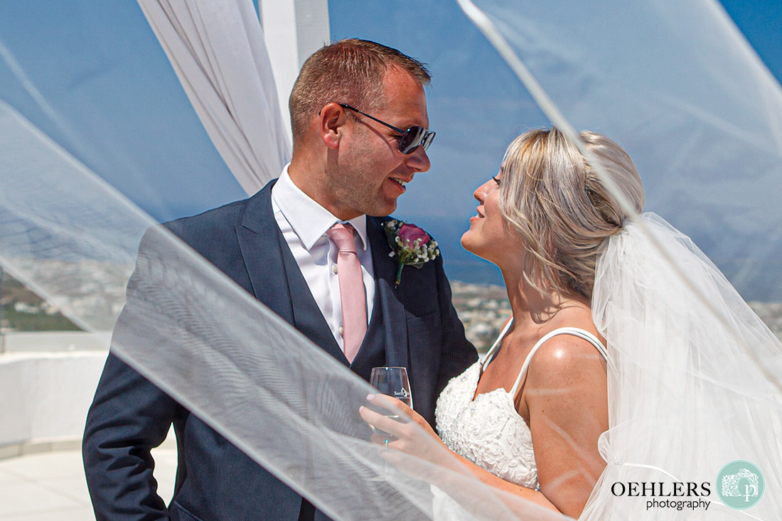Santorini Destination Wedding Photographers - Bride looking at her husband as veil plays in the wind.
