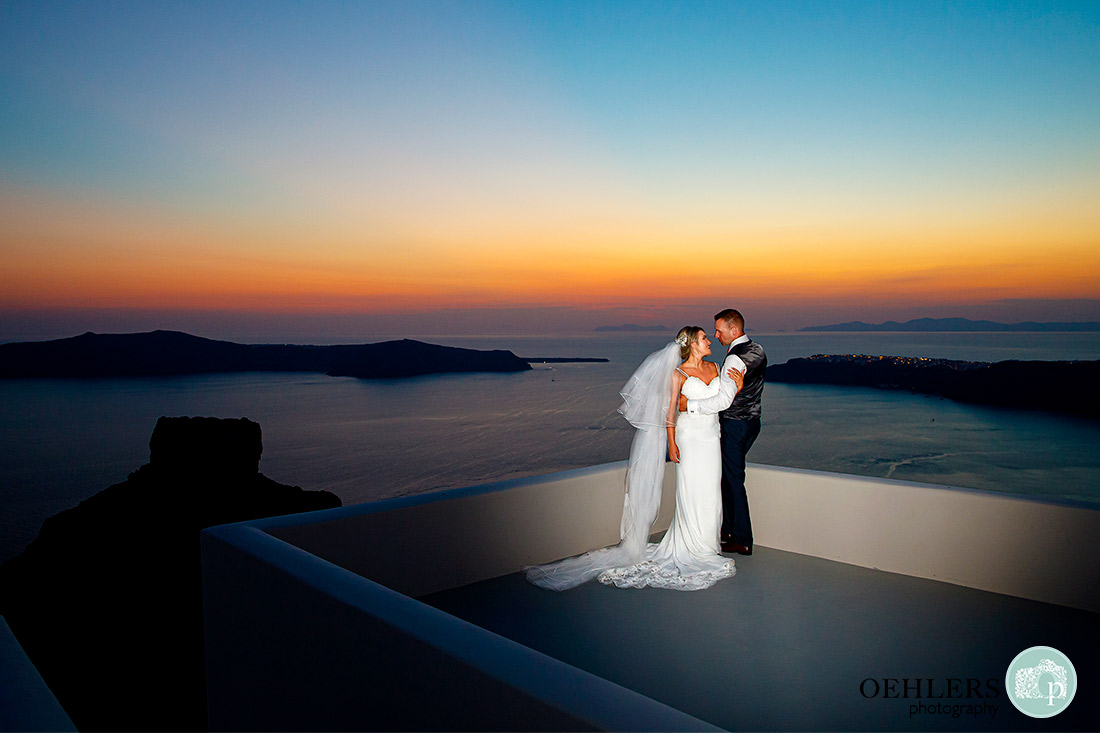 Destination Wedding Photographers - Santorini - bride and groom on a rooftop overlooking the sea with a beautiful sunset.