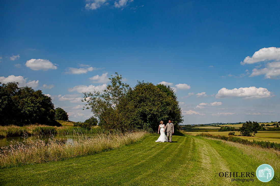 Big blue sky, lake to the left as the bride and groom walk hand in hand towards the camera in the beautiful Leicestershire countryside.